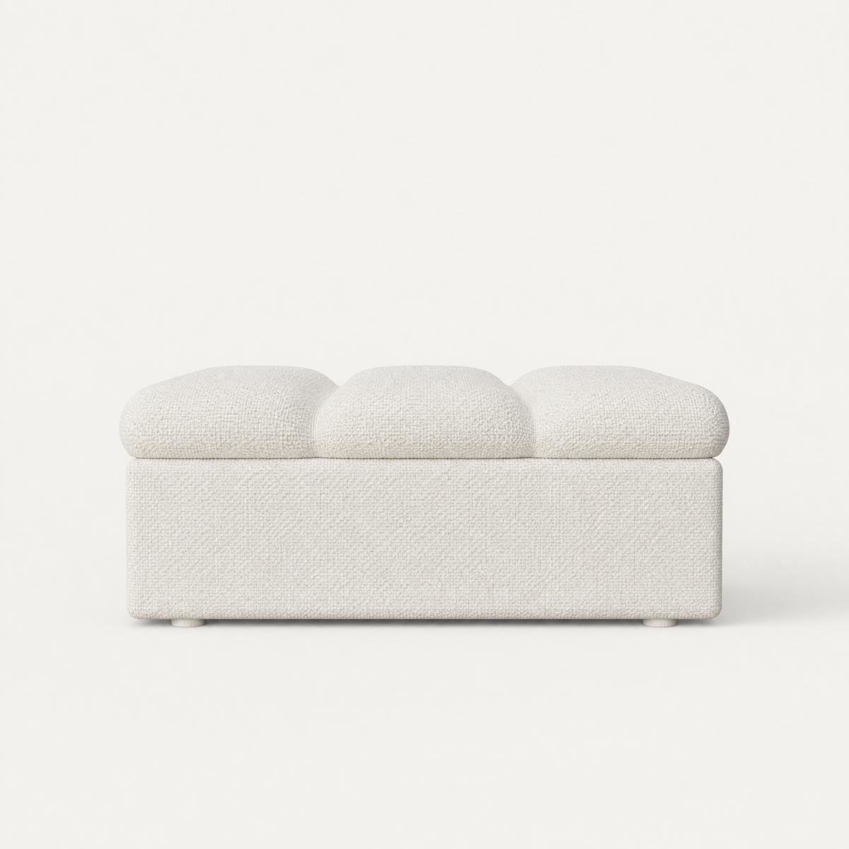 A modern upholstered ottoman with a textured fabric in off-white, featuring a three-segmented top cushion and small round feet, shown against a solid off-white background.