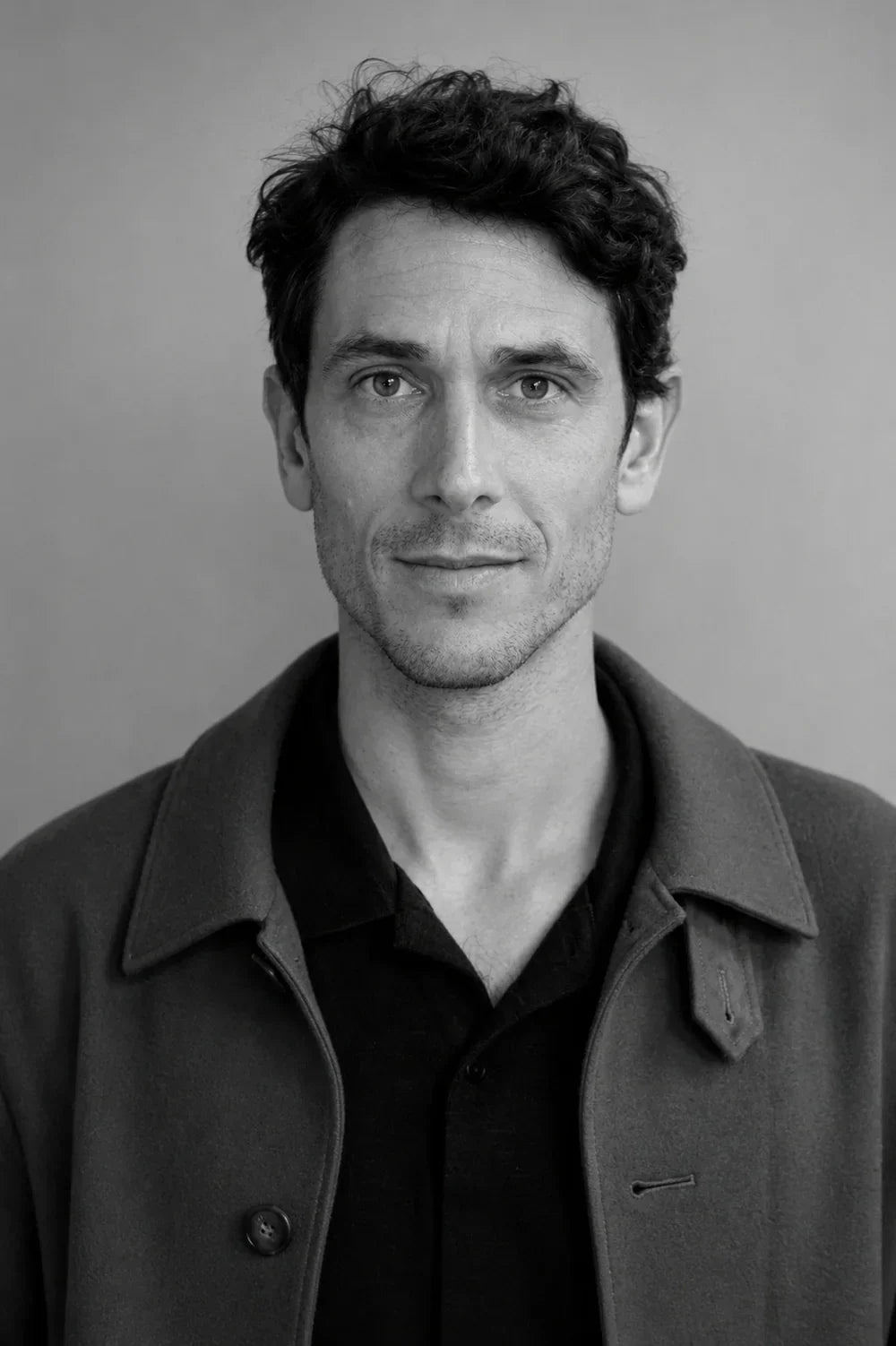 Black and white portrait of a man with curly hair wearing a dark shirt and a grey coat.