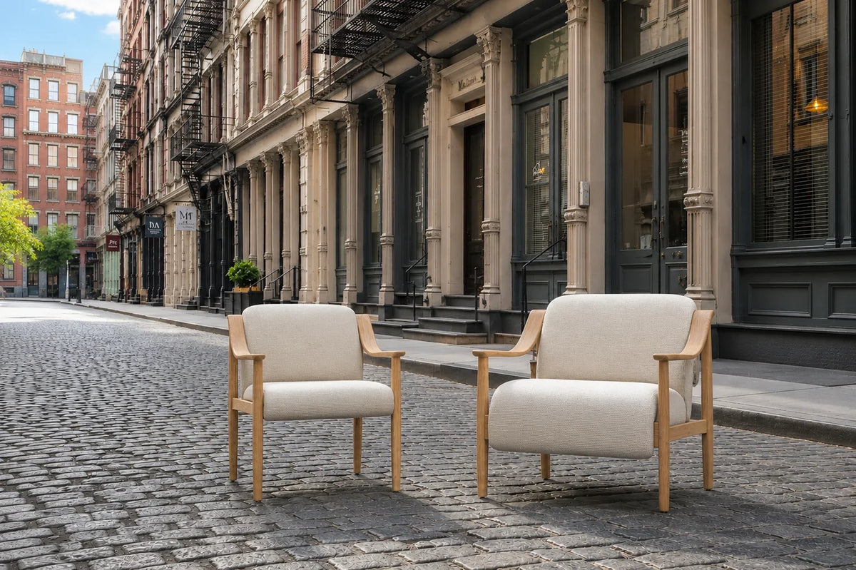 Two modern beige upholstered armchairs with light wood frames on a cobblestone city street.