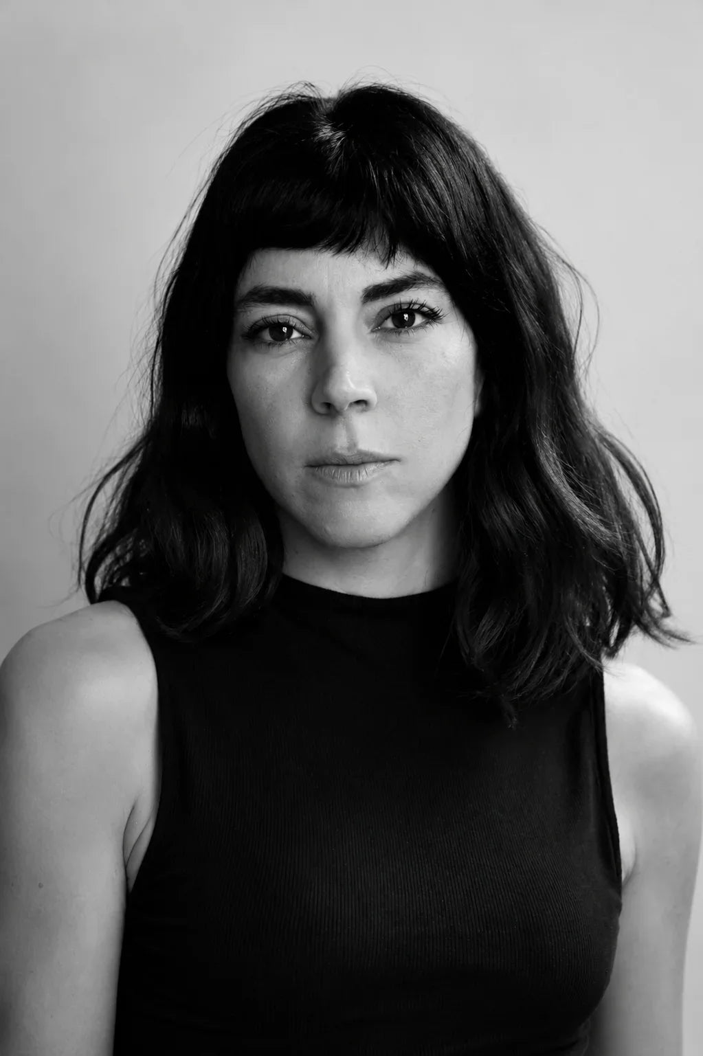 Black and white portrait of a woman with dark wavy hair and bangs wearing a black sleeveless top.