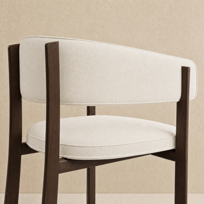 A modern accent chair with a curved backrest and seat cushion upholstered in a light beige textured fabric, supported by a dark brown wooden frame, shown against a neutral beige textured wall.