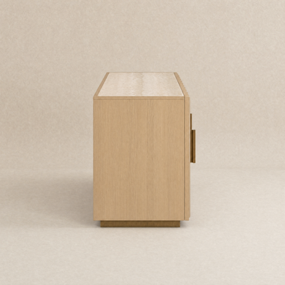 A light oak wood sideboard cabinet shown from a side profile view, featuring a single brass pull handle and a matching brass base, set against a neutral beige textured background.